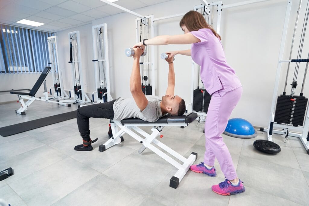 PT practice session showing a physical therapist assisting a patient with a dumbbell chest press on a bench.