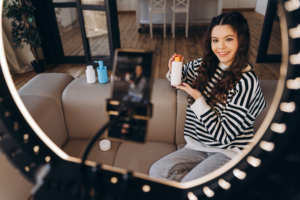 Young teenager vlogger holding cream, showing cosmetics products, recording video